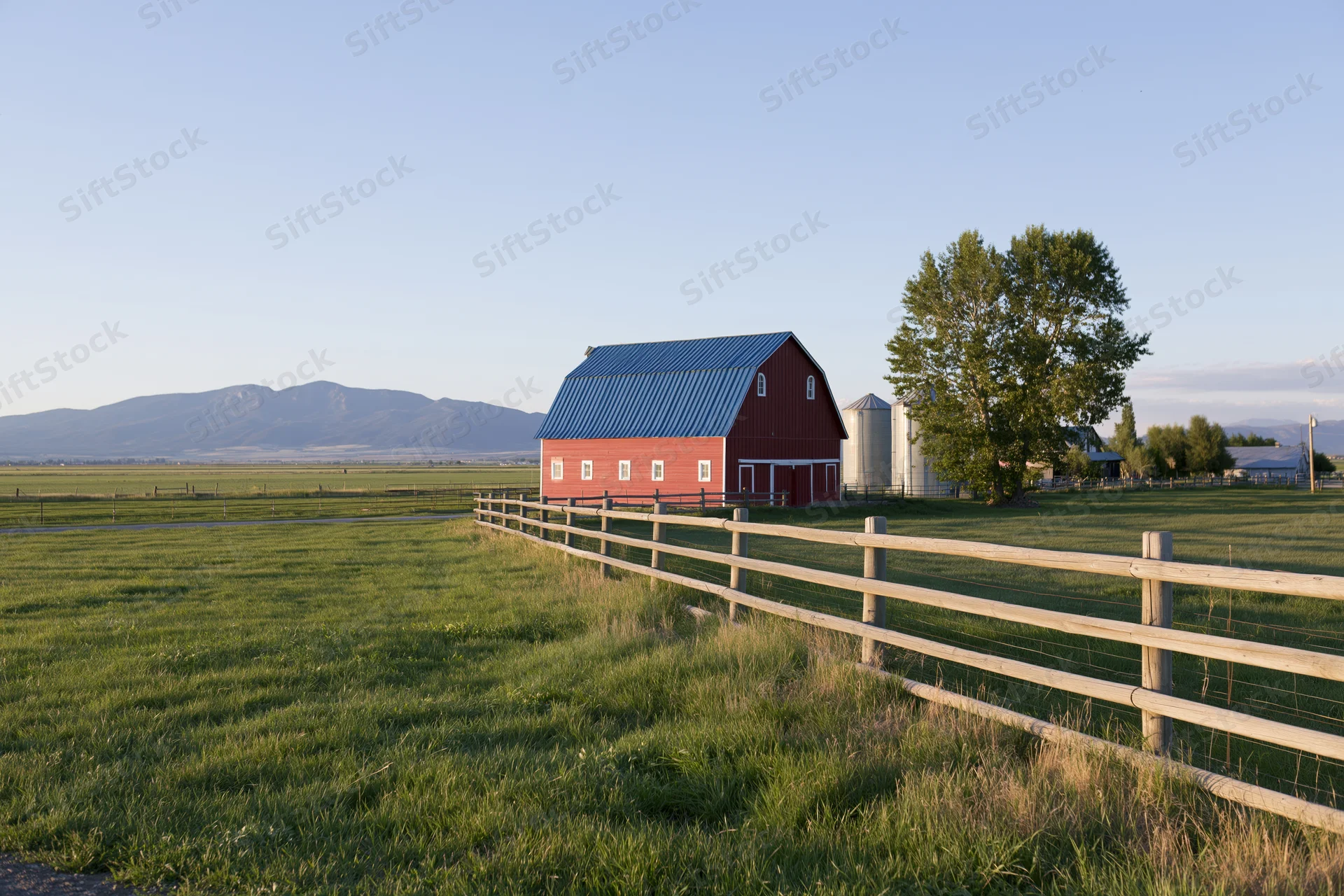 Red barn with blue metal roof in a grassy field bordered by wooden fence near mountains