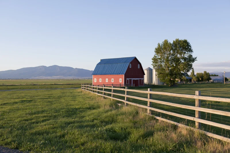 Red barn with blue metal roof in a grassy field bordered by wooden fence near mountains