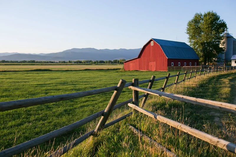 Red barn with blue metal roof in grassy field bordered by wooden fence with mountains in background