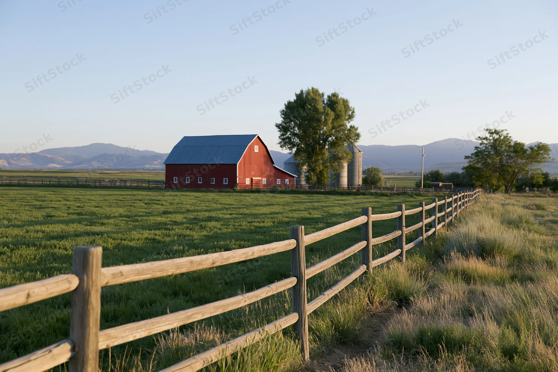 Red barn with blue metal roof in a grassy field bordered by wooden fence and distant mountains