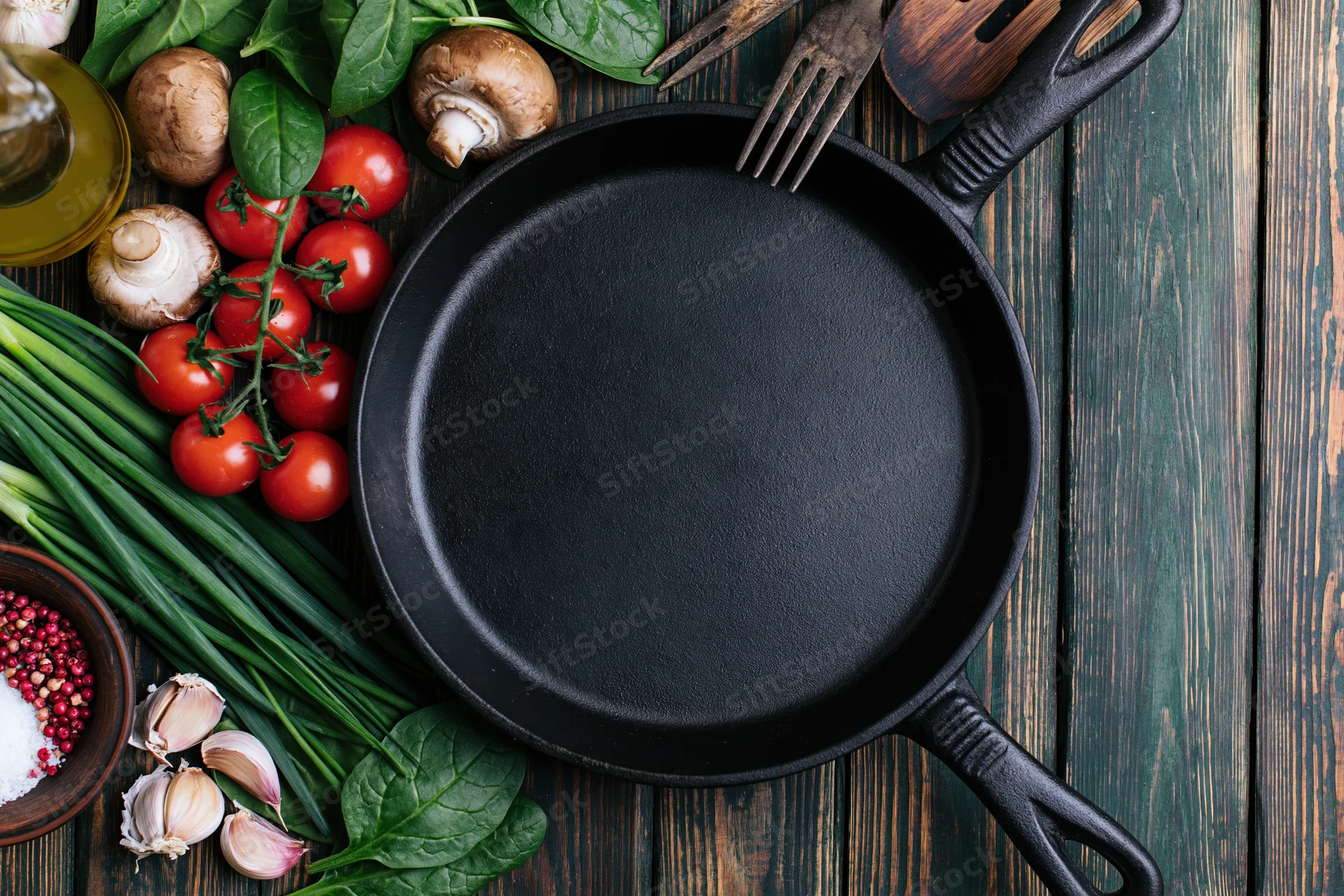 Empty cast iron skillet surrounded by fresh vegetables and herbs on a dark wooden surface