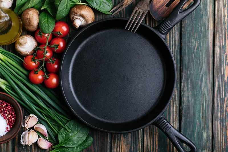 Empty cast iron skillet surrounded by fresh vegetables and herbs on a dark wooden surface