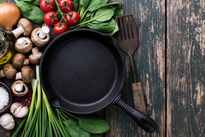 Empty cast iron skillet surrounded by fresh vegetables and cooking ingredients on a rustic wooden table