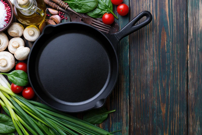 Empty cast iron skillet surrounded by fresh vegetables and cooking ingredients on dark wooden surface