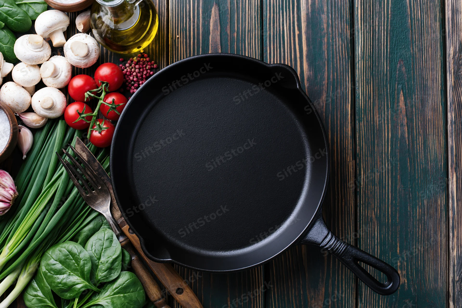 Empty cast iron skillet surrounded by fresh vegetables and cooking ingredients on dark wooden table