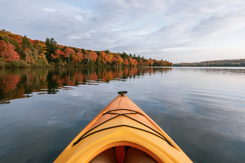 First-person view from the bow of a yellow-orange kayak on a calm lake surrounded by autumn foliage reflecting in the water