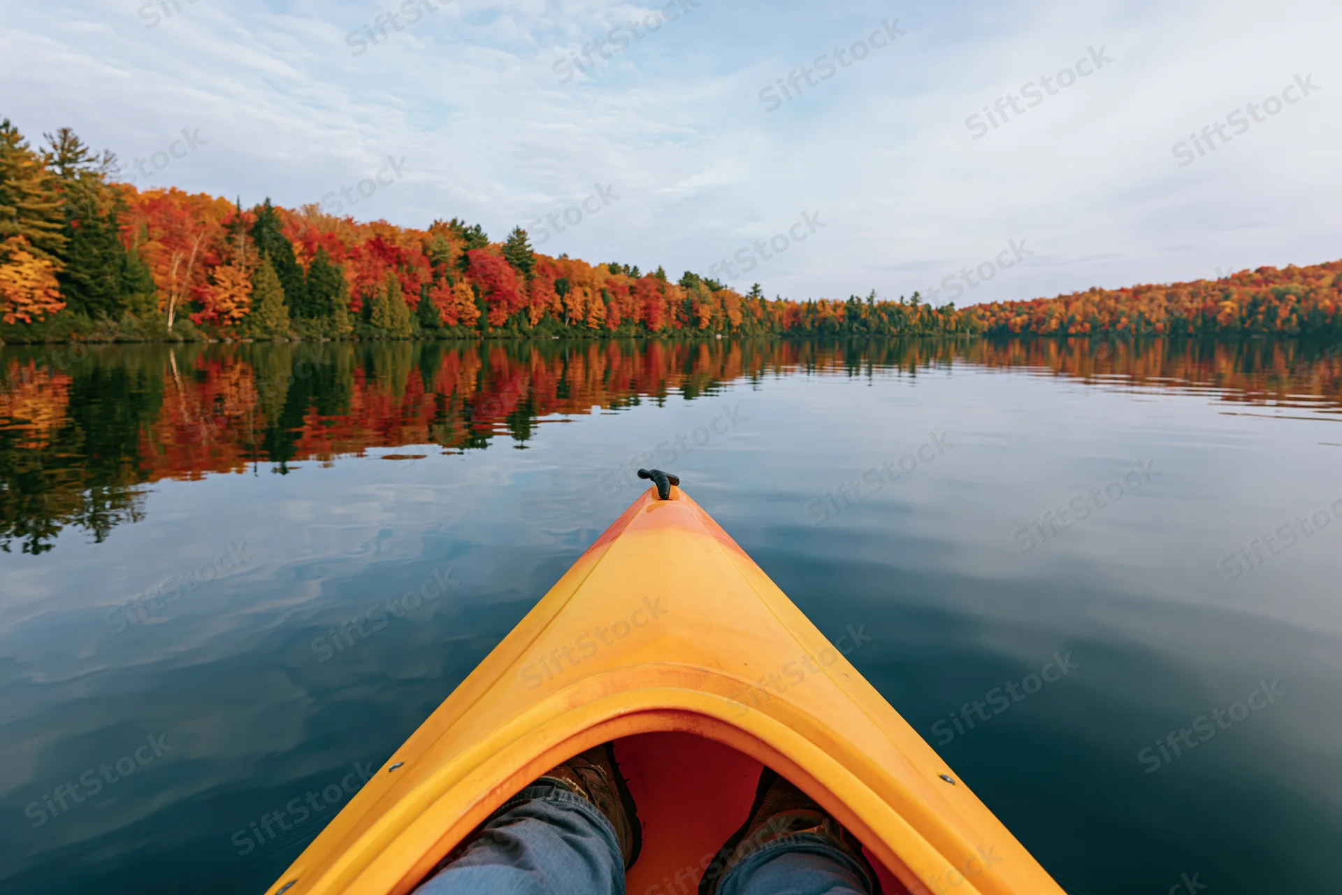 First-person view from bow of yellow-orange kayak on calm lake surrounded by autumn forest reflecting in water