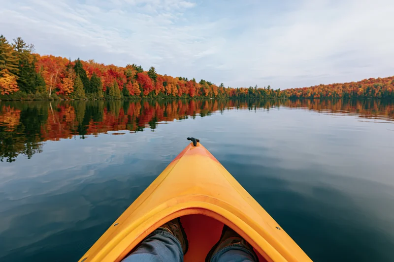 First-person view from bow of yellow-orange kayak on calm lake surrounded by autumn forest reflecting in water