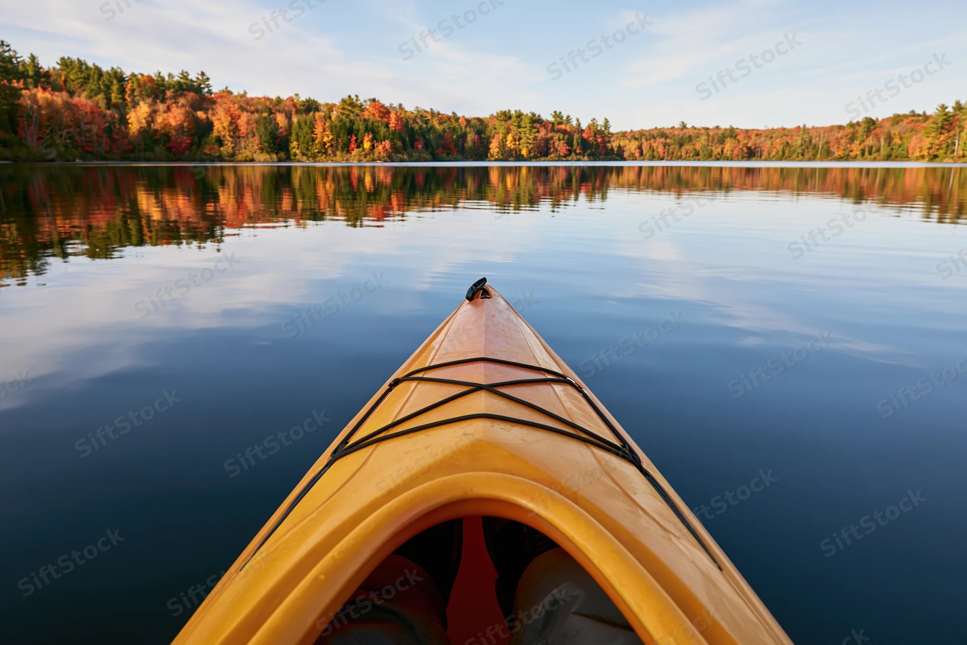 First-person view from the bow of a yellow-orange kayak on a calm lake surrounded by autumn forest reflecting in the water
