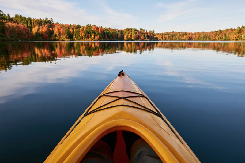 First-person view from the bow of a yellow-orange kayak on a calm lake surrounded by autumn forest reflecting in the water