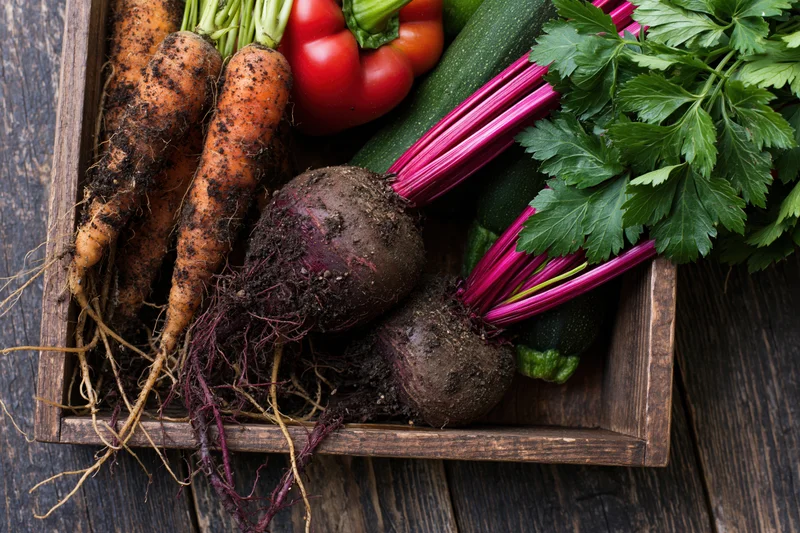 Freshly harvested vegetables with soil still on roots arranged in a wooden crate on a dark wood surface