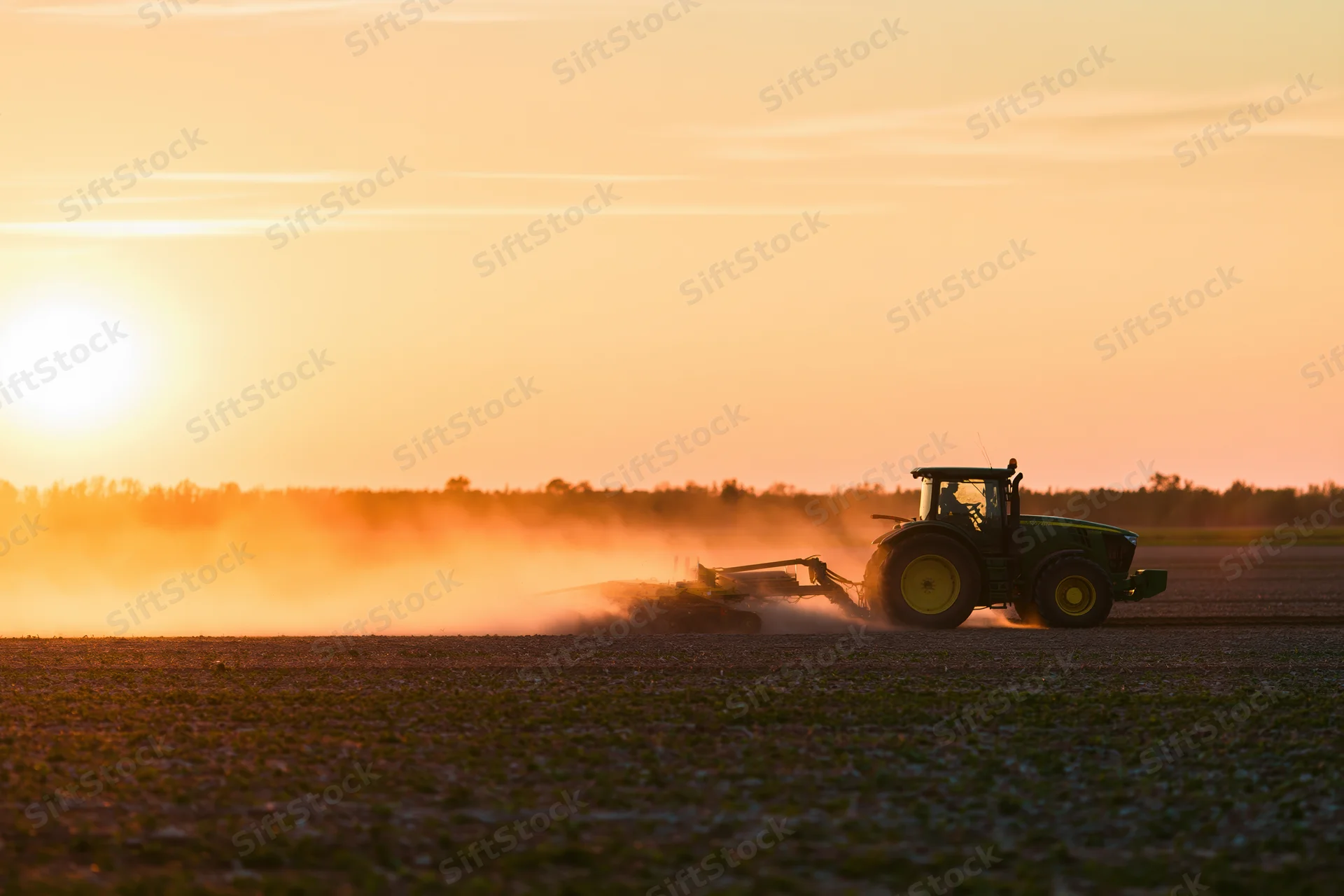 Green tractor pulling tillage equipment across a flat agricultural field at sunset