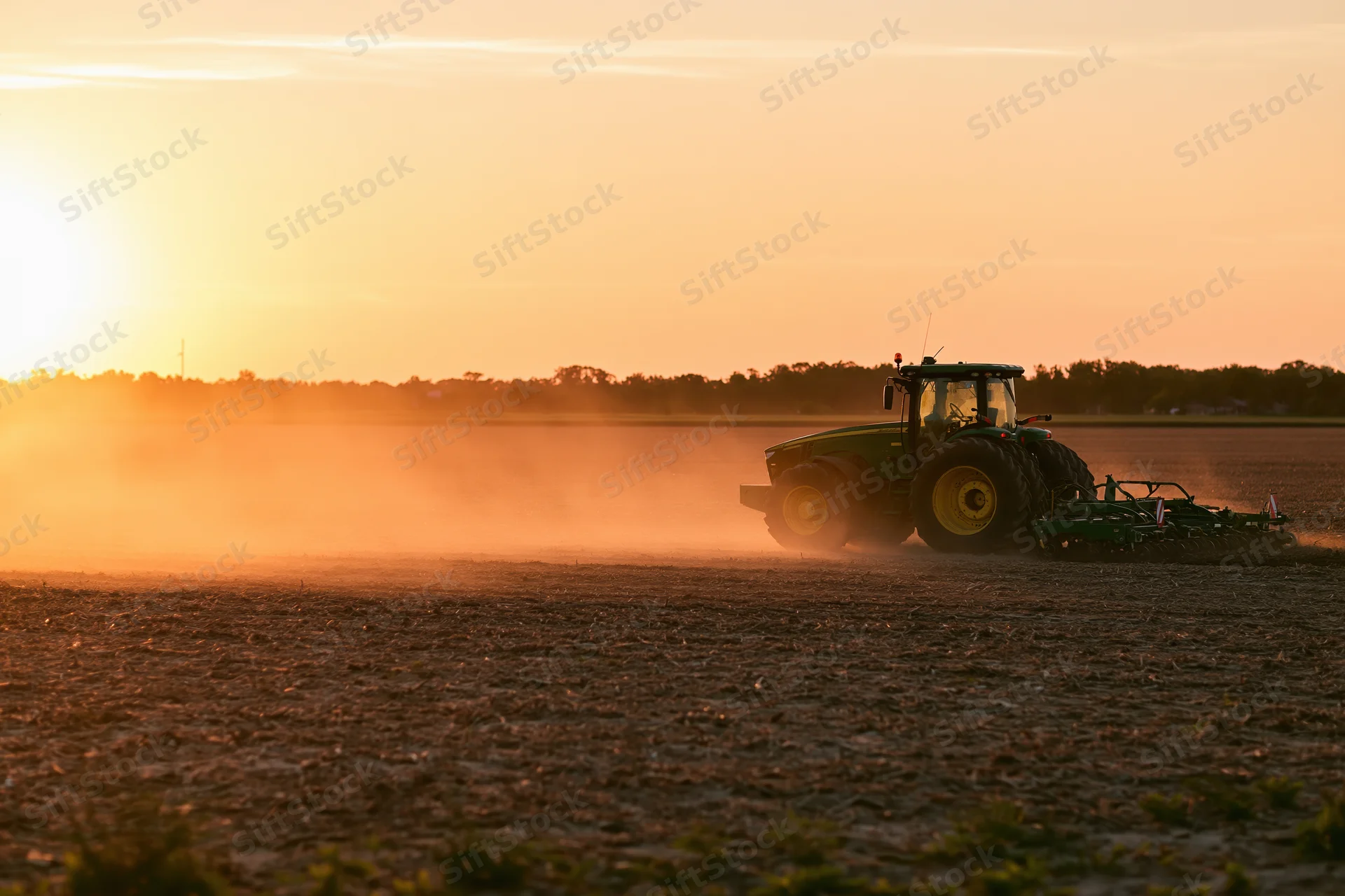 Green tractor pulling tillage equipment across a flat field at sunset