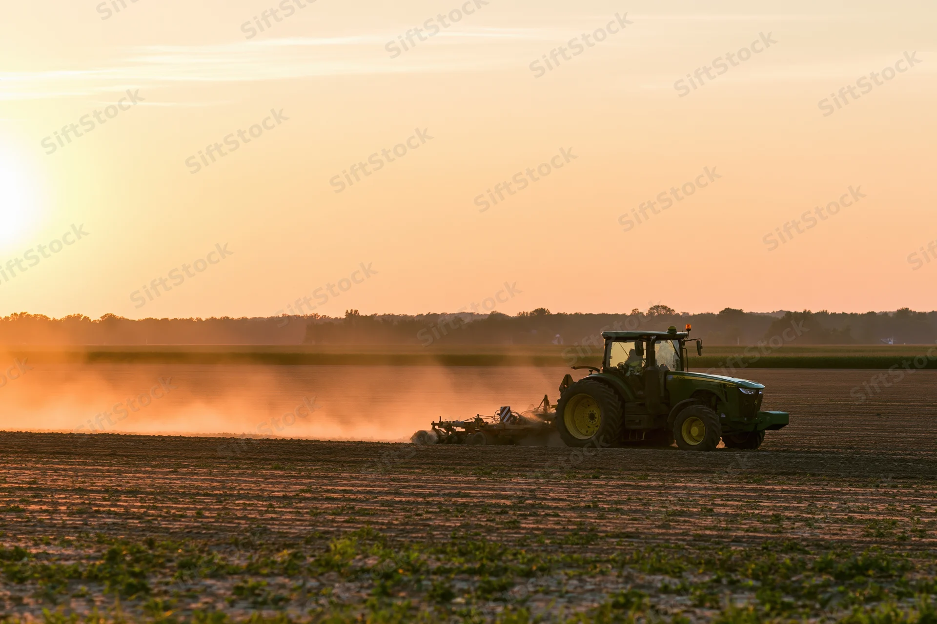 Green tractor pulling tillage equipment across a flat agricultural field at sunset