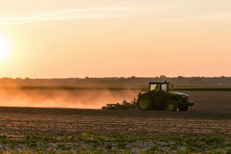 Green tractor pulling tillage equipment across a flat agricultural field at sunset