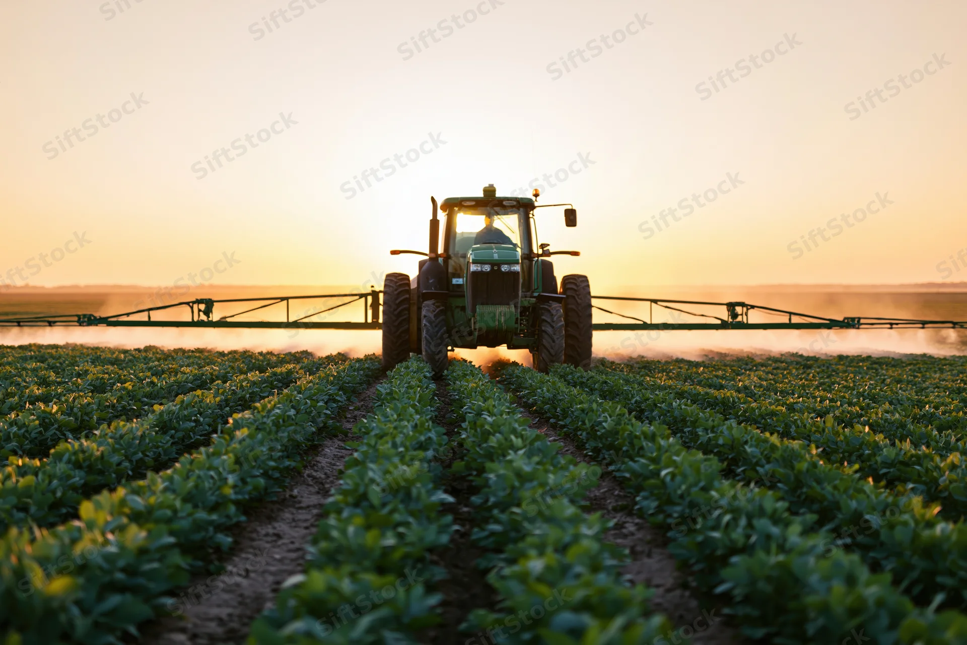 Green tractor with extended spray boom applying pesticide in a field at sunset