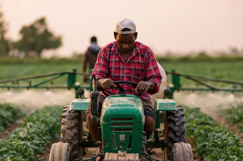 Indian farmer operating small green tractor with spray boom in crop field