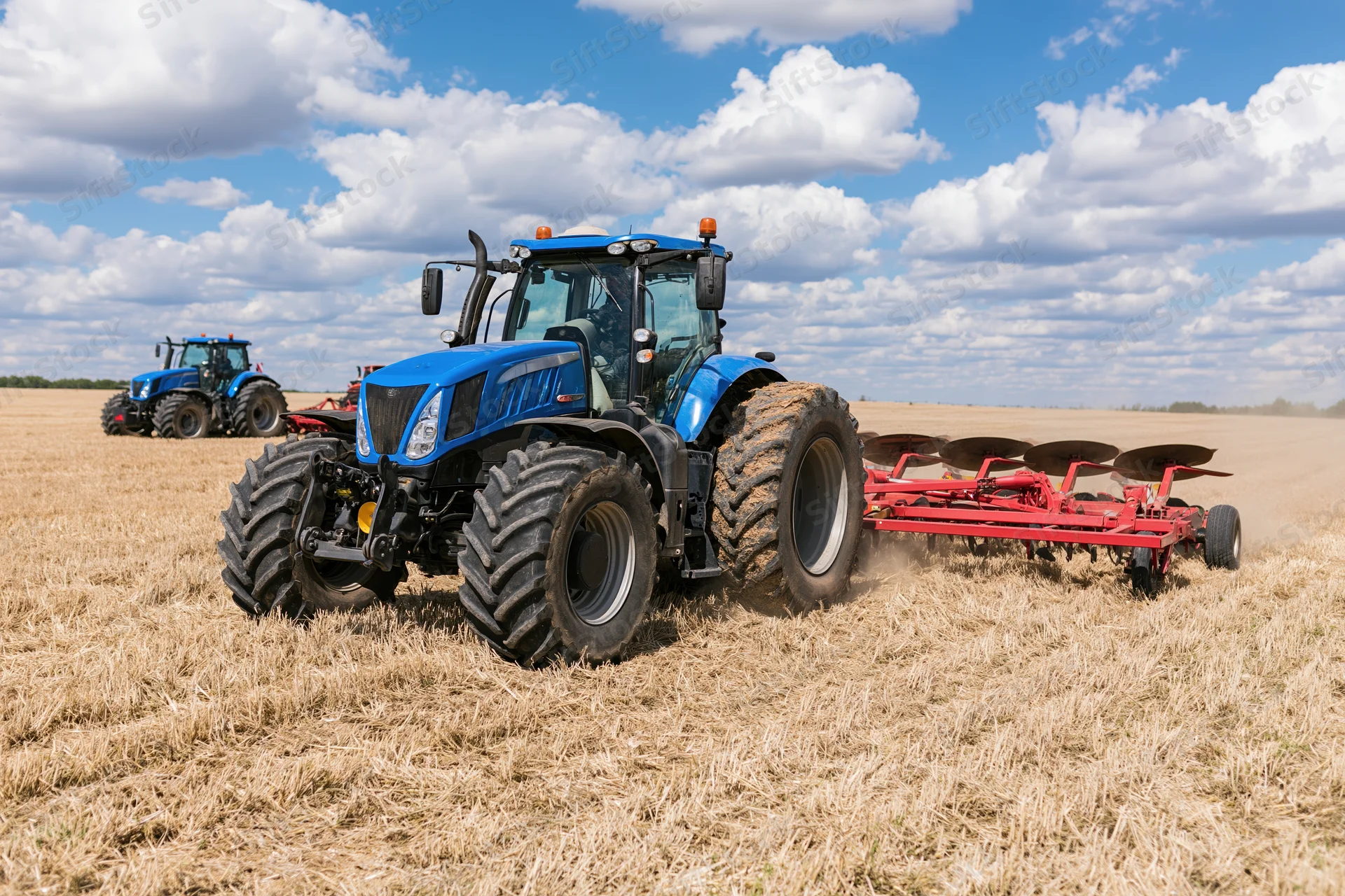 Blue tractor pulling red disc harrow across harvested field under blue sky with clouds
