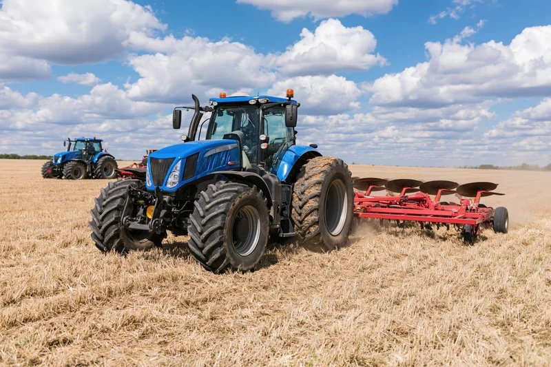 Blue tractor pulling red disc harrow across harvested field under blue sky with clouds