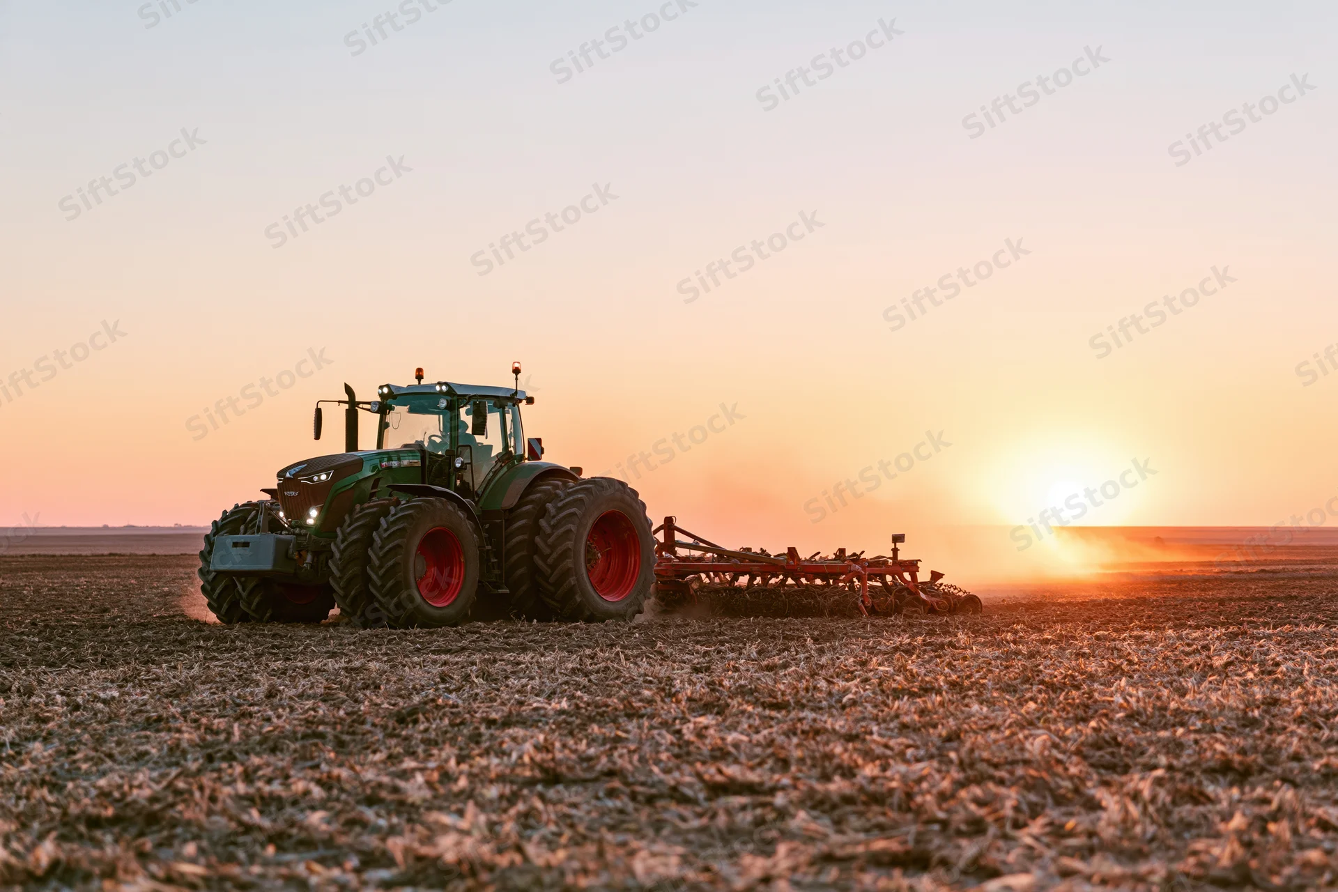 Green tractor pulling a cultivator across a harvested field at sunset