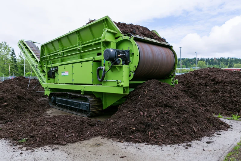Green industrial compost screening machine processing organic material outdoors with mulch piles around it