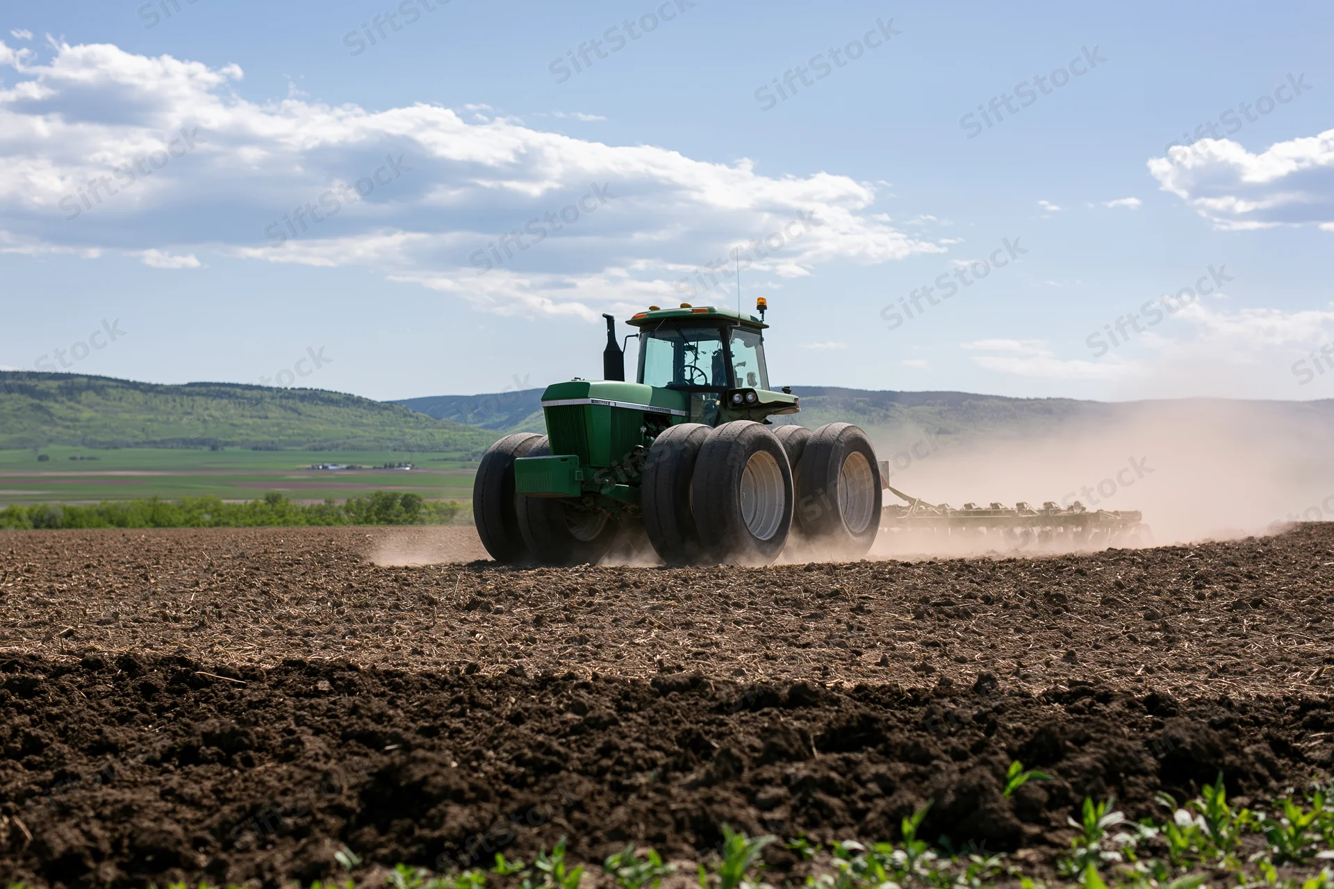 Green tractor pulling seeder across tilled field under partly cloudy sky
