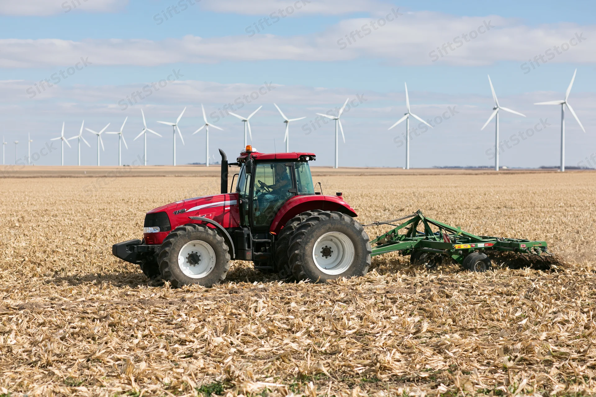 Red tractor pulling green tillage implement across harvested cornfield with wind turbines in background