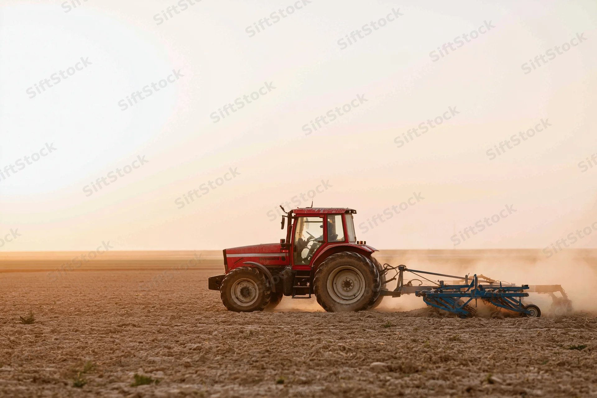 Red tractor pulling a soil cultivator across a flat agricultural field at sunset