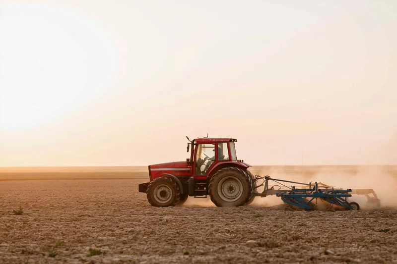 Red tractor pulling a soil cultivator across a flat agricultural field at sunset