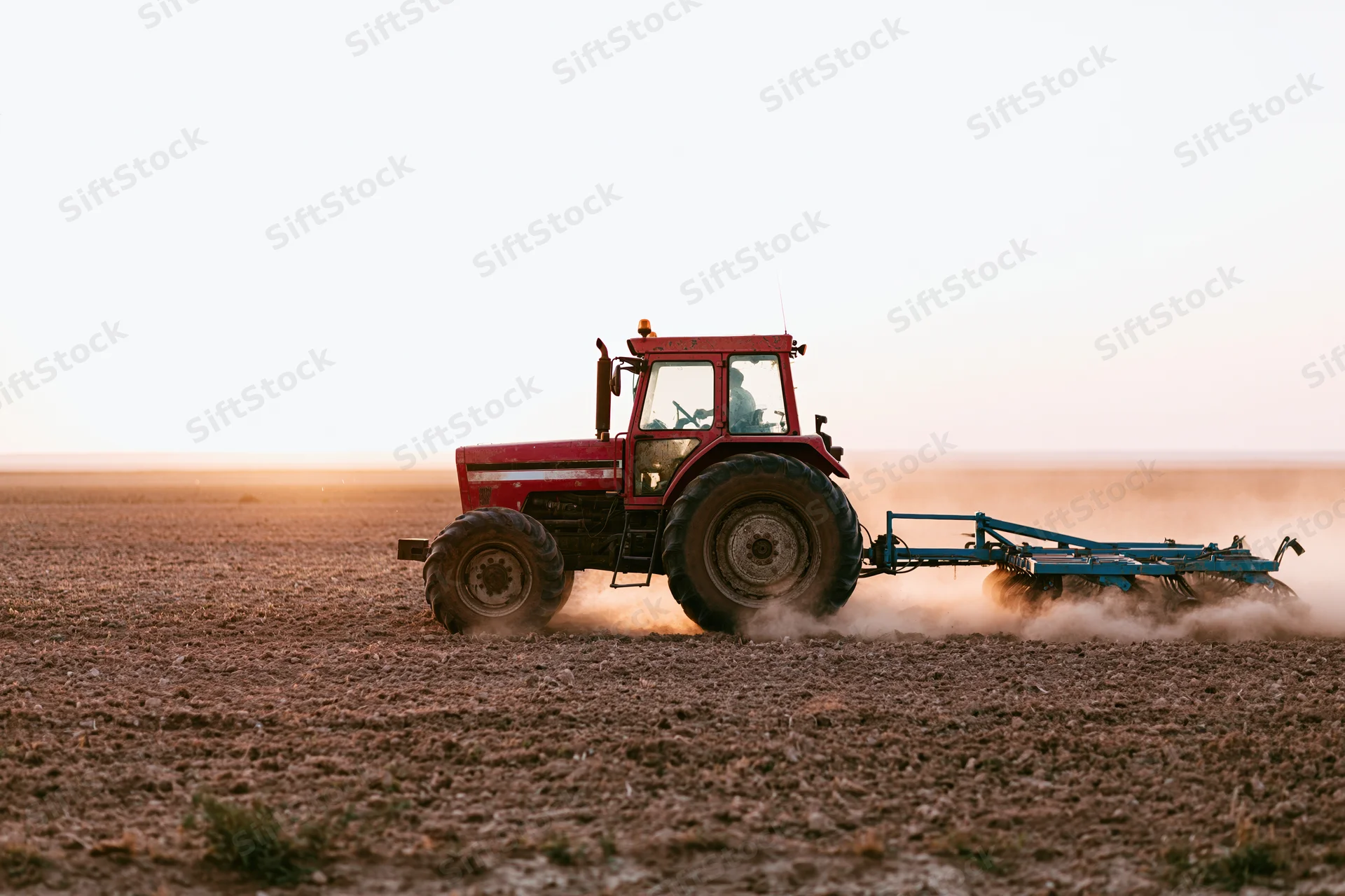 Red tractor pulling a blue soil cultivator across a flat agricultural field