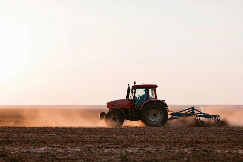 Red tractor pulling a soil cultivator across a flat agricultural field at sunset