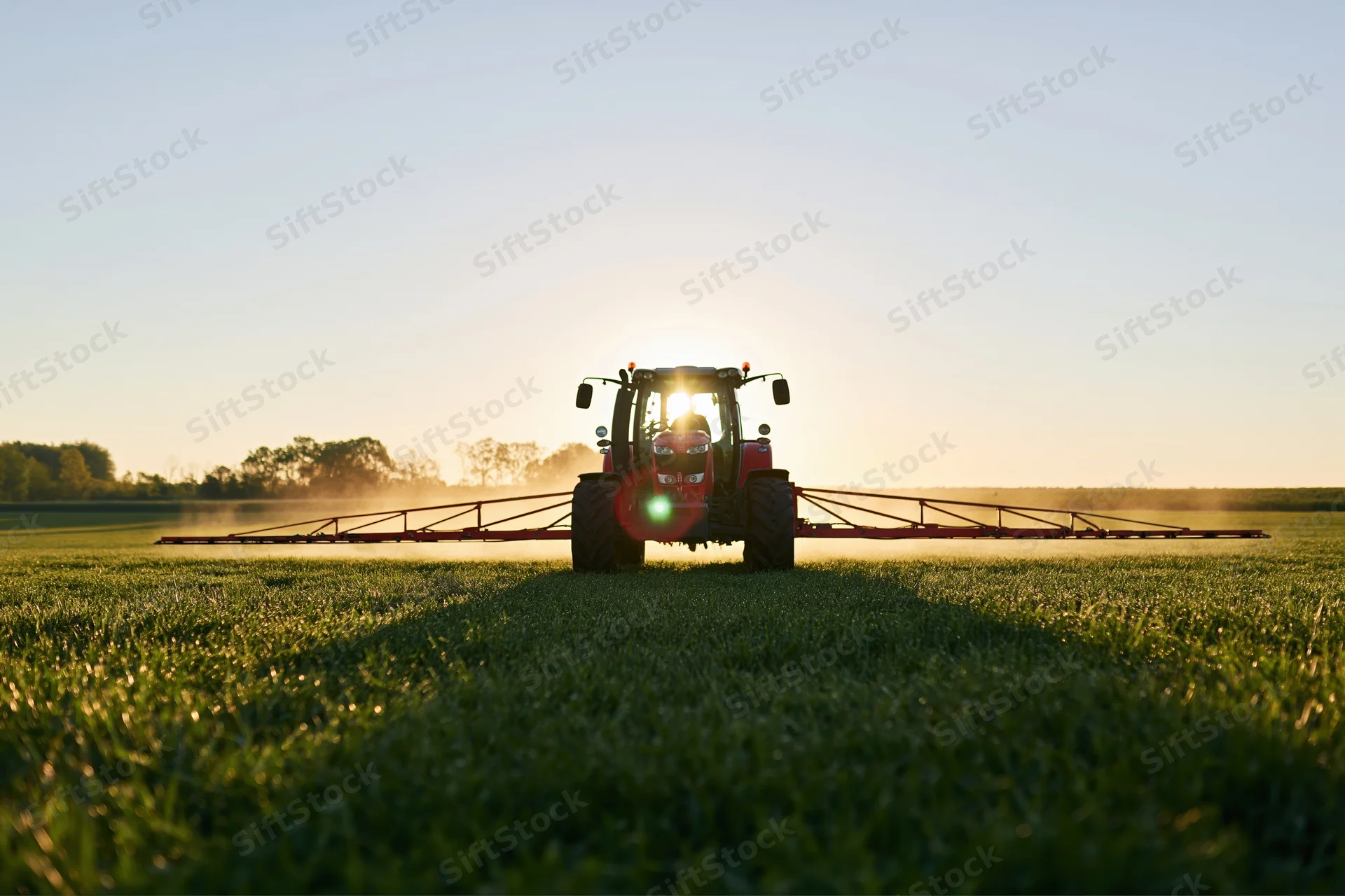 Red tractor pulling wide sprayer across flat green field at sunset