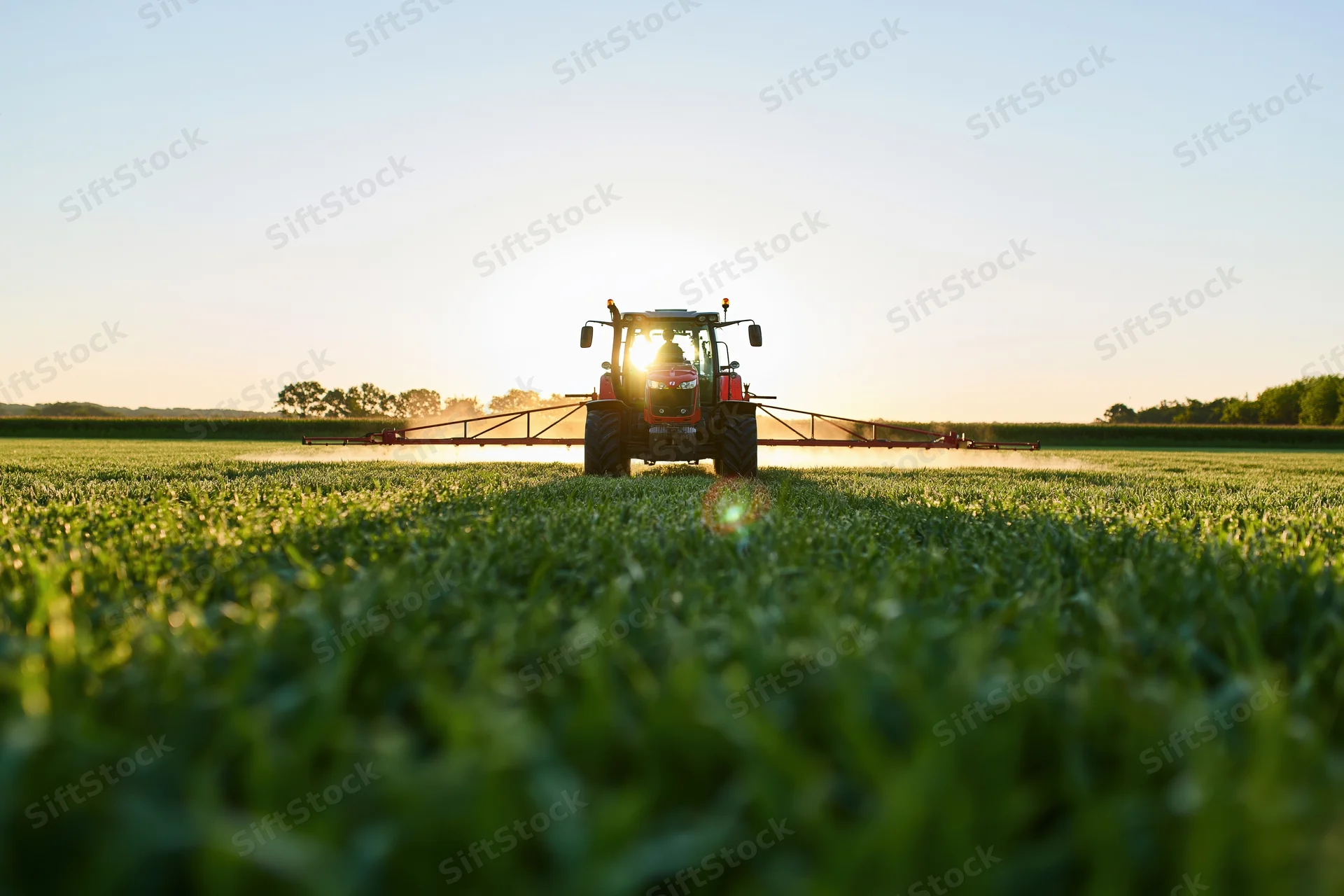 Red tractor pulling wide sprayer across flat green field at sunset