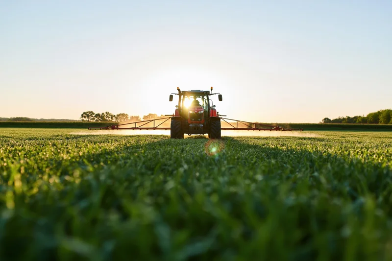 Red tractor pulling wide sprayer across flat green field at sunset