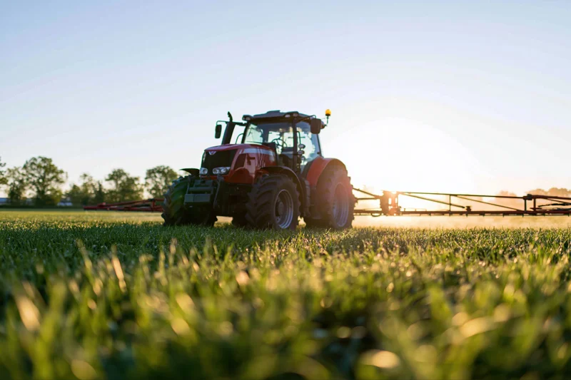 Red tractor pulling a wide sprayer across a flat green field at sunrise