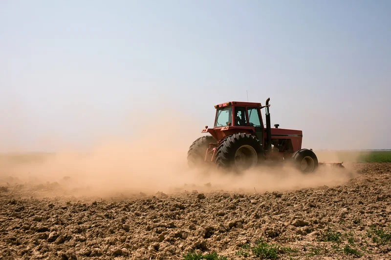 Red tractor tilling a dry field kicking up a large dust cloud