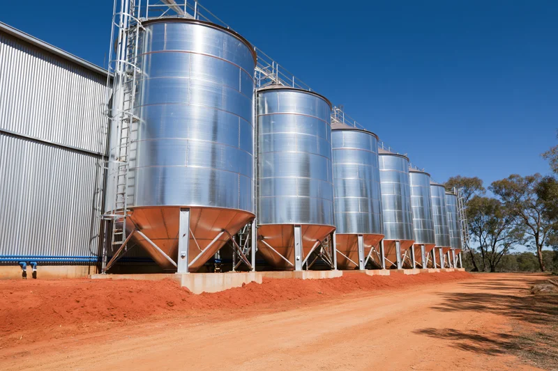 Row of galvanized steel grain silos with conical bases aligned along a red dirt road beside a corrugated metal building under a clear blue sky