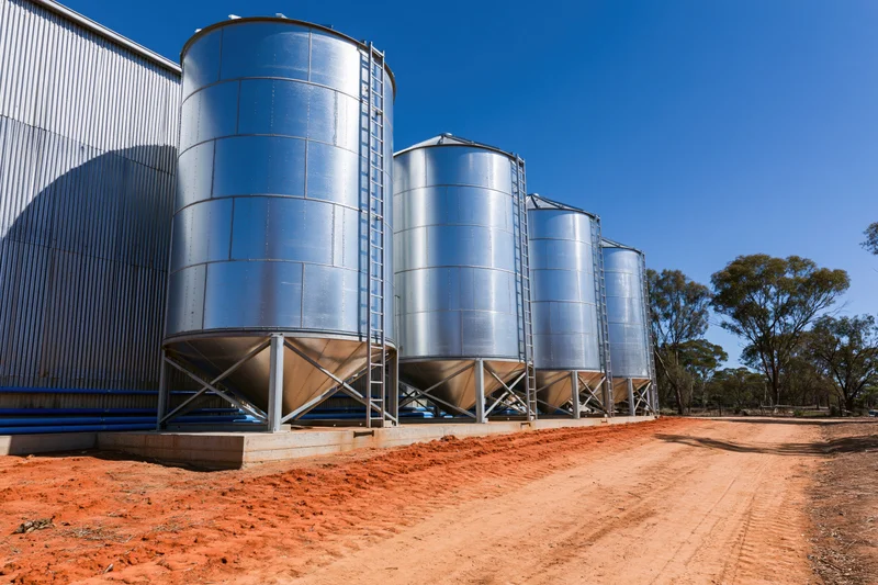 Row of galvanized steel grain silos with conical bases on a red dirt farm site