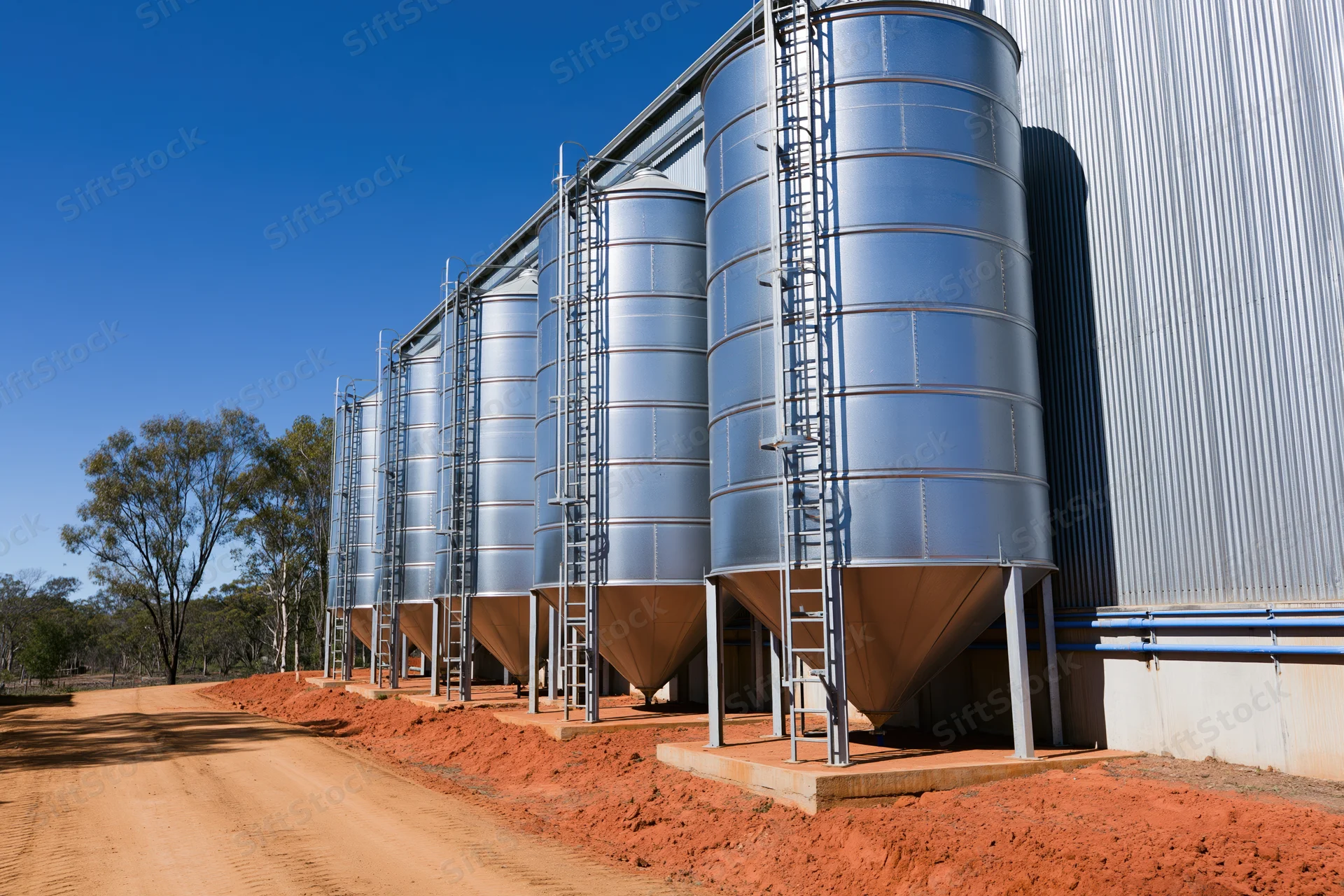 Row of galvanized steel grain silos with conical bases aligned along a dirt path in a rural farm setting