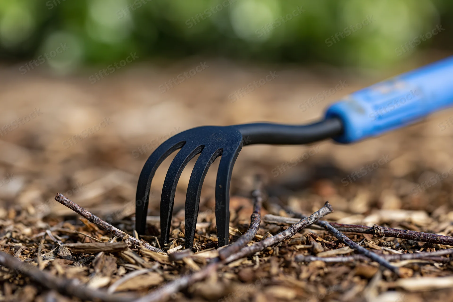 Small hand cultivator with blue rubber grip resting on dry mulch and twigs in garden