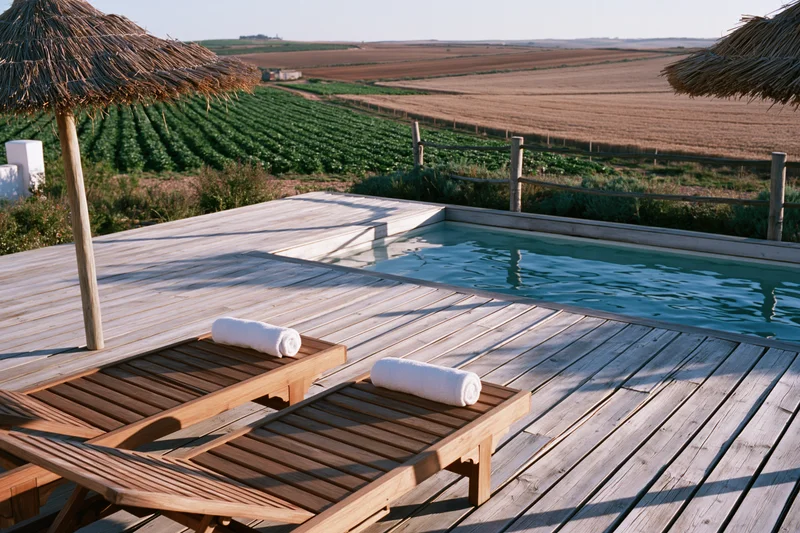 Small rectangular swimming pool set into a weathered wooden deck with lounge chairs and thatched umbrellas overlooking farmland