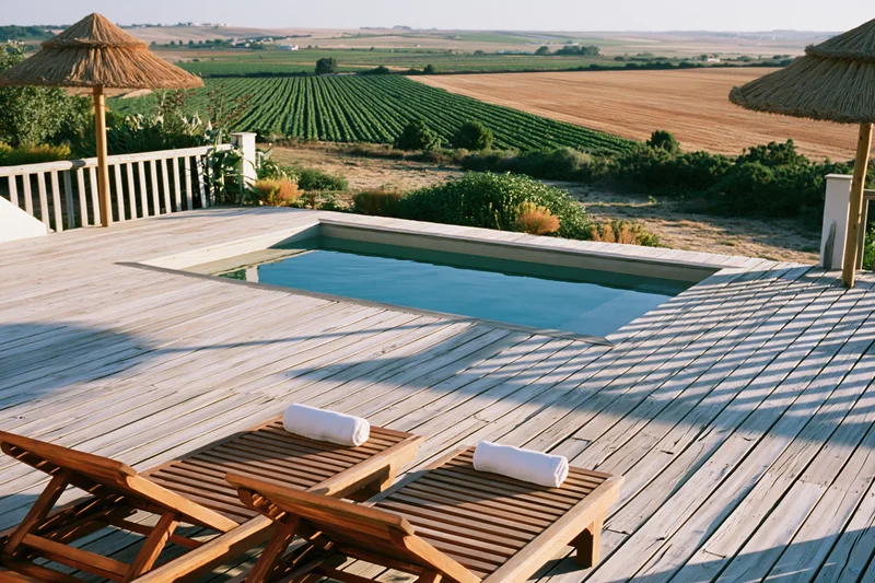 Small rectangular swimming pool set into a weathered wooden deck with lounge chairs and straw umbrellas overlooking vineyards and farmland