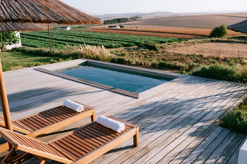 Small rectangular swimming pool set into a weathered wooden deck with lounge chairs and towels, overlooking rural farmland fields under a clear sky