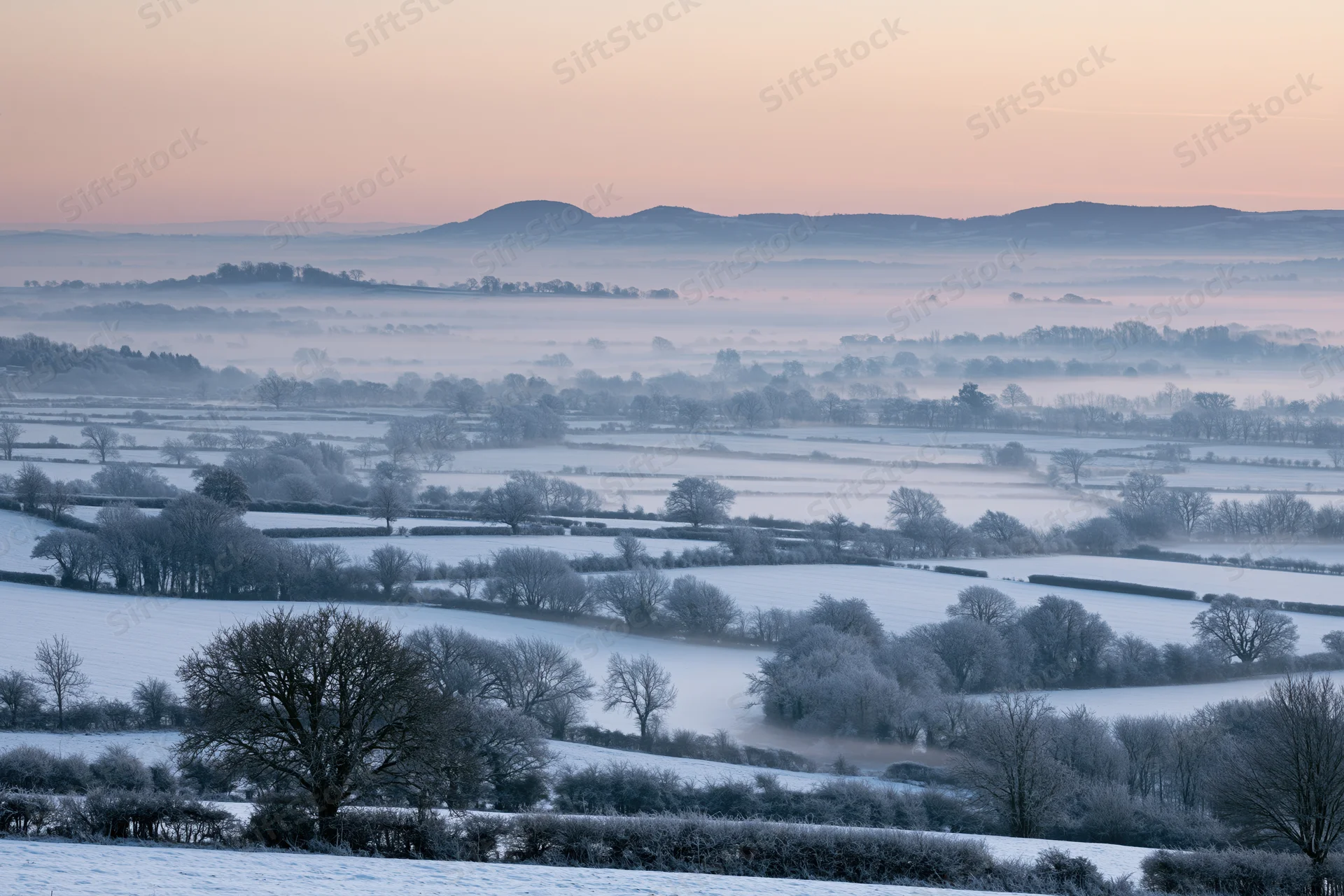Snow-covered english countryside valley at dawn with frosty trees and misty hills in the distance