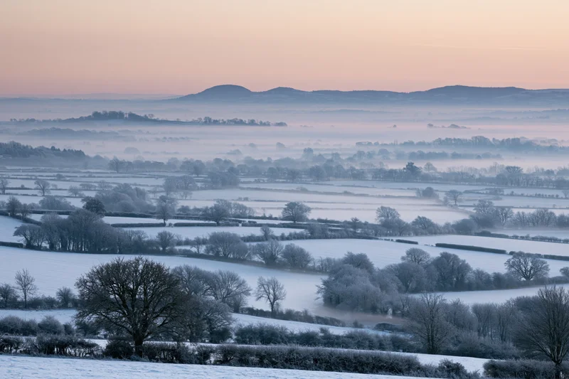 Snow-covered english countryside valley at dawn with frosty trees and misty hills in the distance