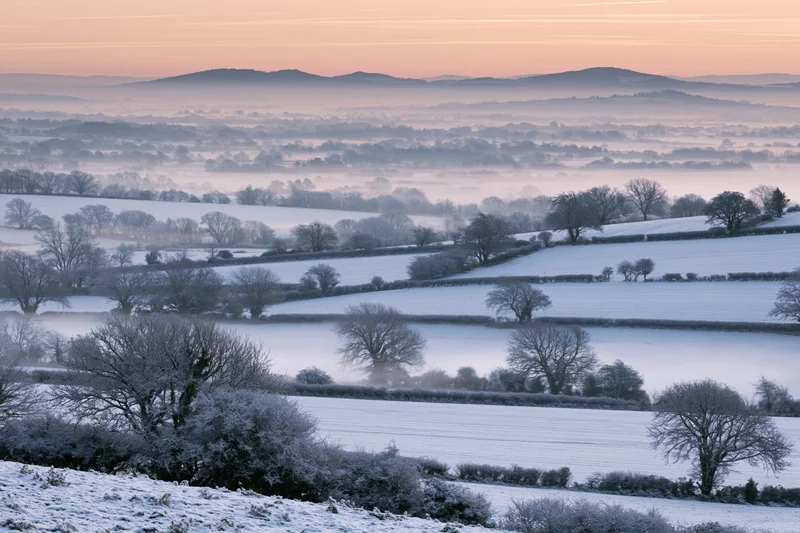 Snow-covered english countryside valley at dawn with rolling fields and bare trees under a misty sky