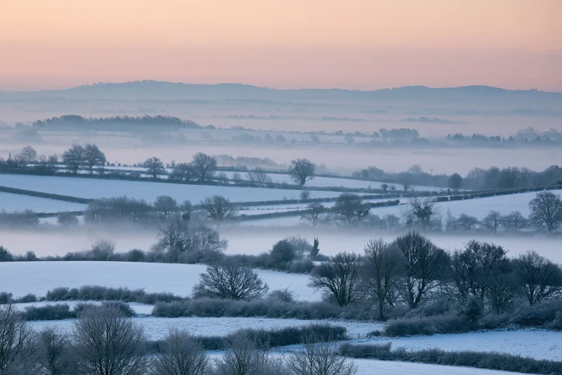 Snow-covered english countryside valley at dawn with misty hills and frosty trees