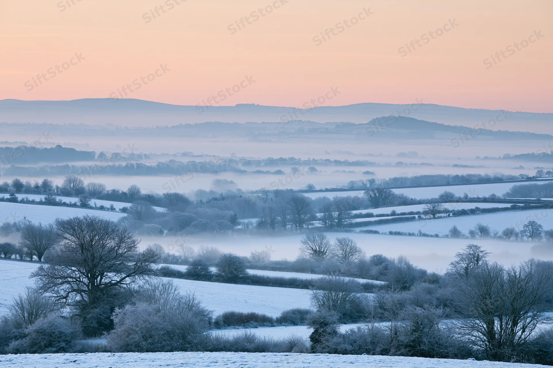 Snow-covered english countryside valley at dawn with misty hills and frosty trees