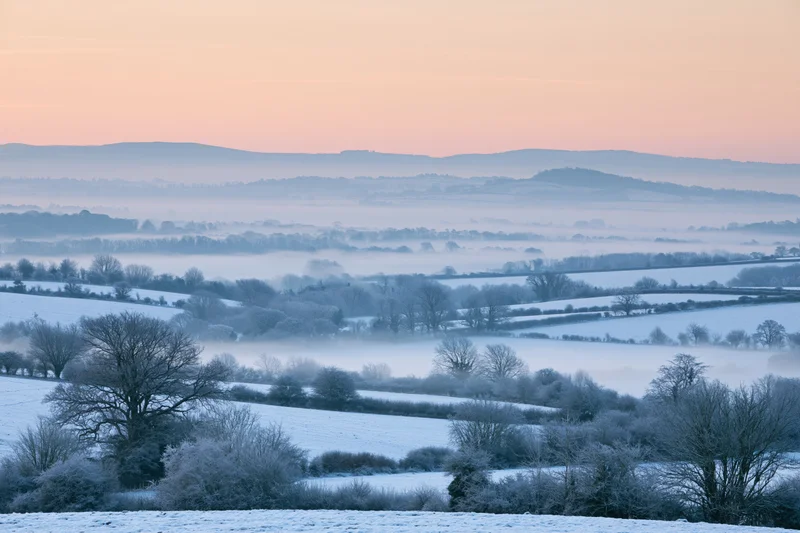 Snow-covered english countryside valley at dawn with misty hills and frosty trees