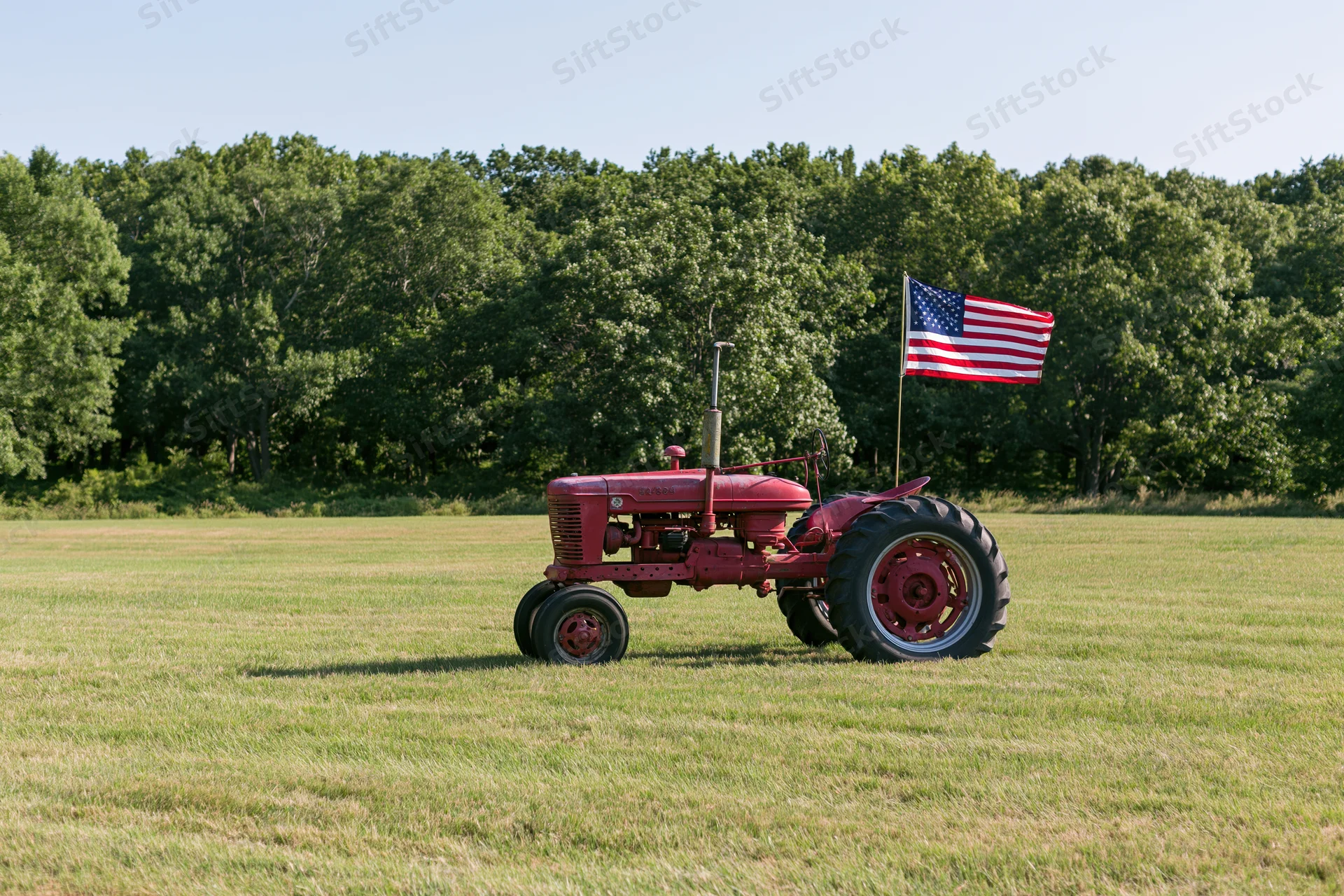 Vintage red farm tractor parked in a mowed grass field with american flag attached, surrounded by trees under clear sky